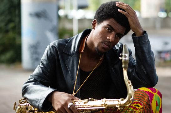 Young man in black jacket holding a saxophone, with hand on head in thoughtful pose.