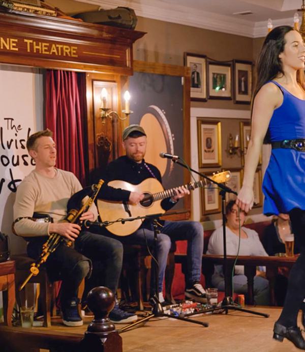 Several musicians play Irish traditional music and a woman in blue dress dances in front.