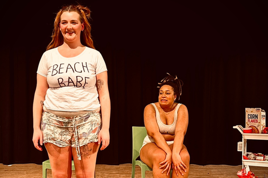 One woman wearing a t shirt that says 'Beach Babe' stands up on the left, while another sits on a chair to her right with her arms on her knees.