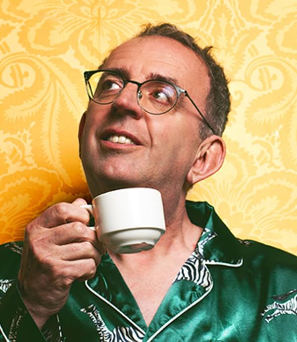 A man in glasses and green pyjamas holds a cup of tea in front of a yellow wall.