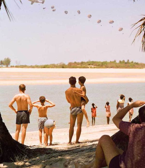 A group of men, women and children on a beach stare into the distance where we can see a plane dropping a row of parachutes.