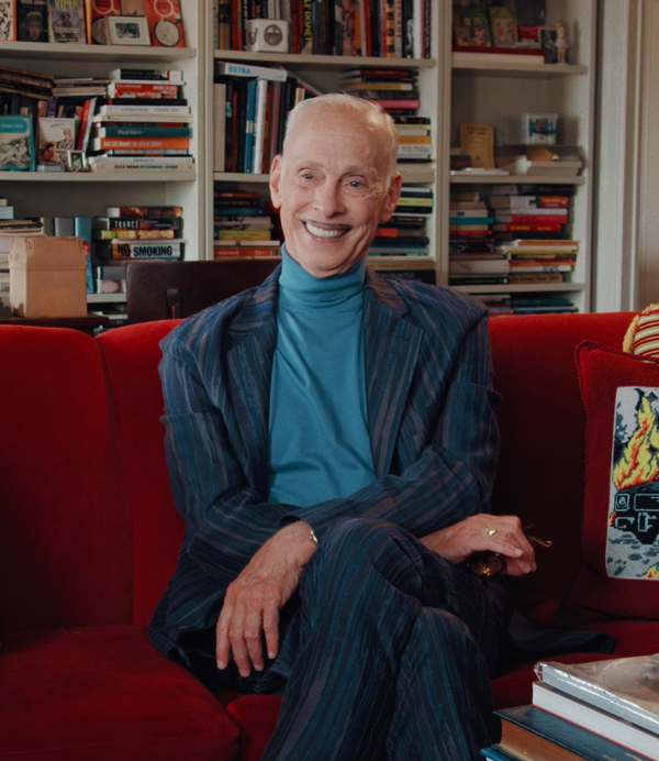 A smiling man (director John Waters) sits on a red sofa in his apartment. He is surrounded by books and one of the cushions has a picture of a burning police car on it.