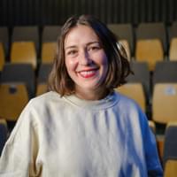 A woman in her late thirties with short brown hair and bright red lipstick on smiles at the camera. Behind her are a collection of flip-up seats in a theatre space.