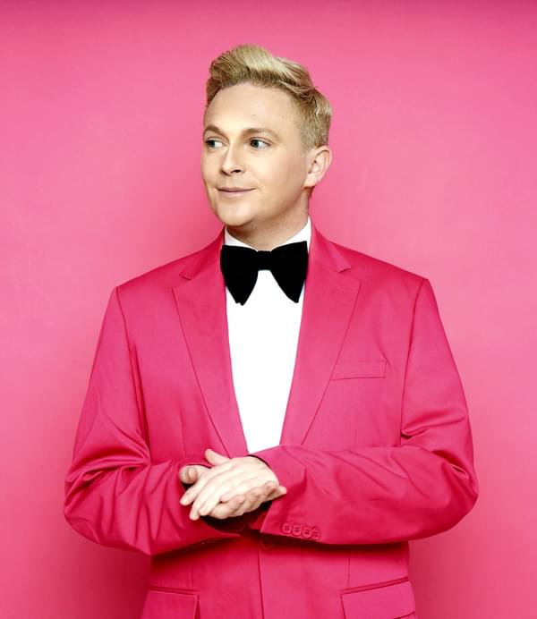 A main in a pink suit and wearing a bow tie in front of a pink background.