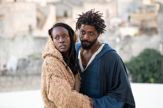 A bearded Black man and a hooded Black woman dressed in cloaks hold each other.