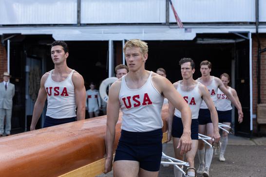 A group of young men in white vests with the red letters USA printed on them carry an orange rowing boat.