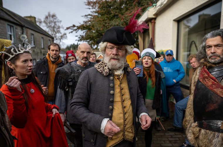 A group of people in a mix of historical costume and everyday clothing gather on a village street. At the centre stands an older man with a white beard and glasses, dressed in an 18th-century-style grey coat, gold waistcoat, and a black tricorn hat adorned with red feathers. To his left, a young woman wears a red medieval-style dress and a decorative antler crown. To his right, a man is dressed in dark fur-trimmed armour with a red sash. Behind them, a crowd of onlookers includes people in both period costume and modern clothing. The setting appears to be a small Scottish town on an overcast day.