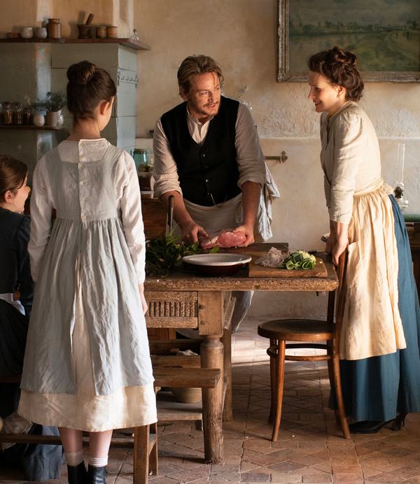 Three women and one man in 19th century dress stand at a kitchen table. The man is preparing a meal, holding a slab of beef.