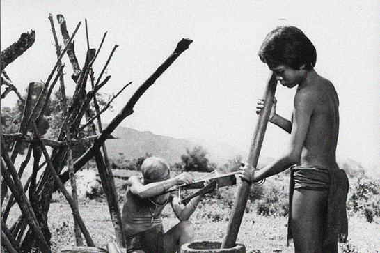A Vietnamese boy wearing a loincloth uses a large pestle & mortar whilst an old man sat behind him works on a crossbow.
