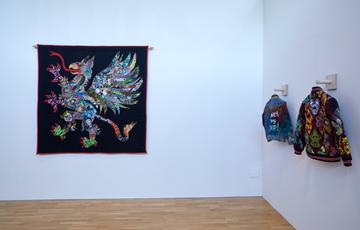 Install shots in an art gallery - big embroidery with black background and a  mythical creature with lots of colours next to two embroidered jackets