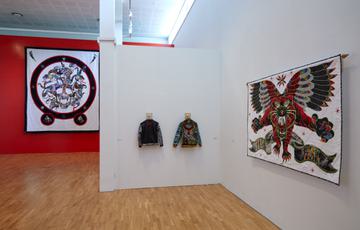 Gallery installation with large red wall and a big embroidered quilted piece, two jackets with embroidery, and a large mythical creature embroidered on a quilt hanging in a gallery