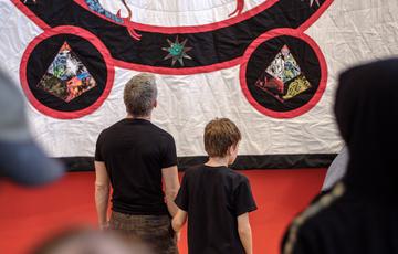 Two people stand in an art gallery facing a large quilted embroidered artwork