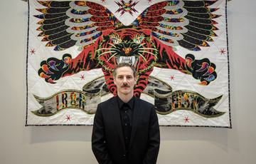 Ben Venom stands in front of a quilted artwork hanging in a gallery