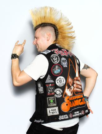 White man with a mohawk faces right with his back to the camera making the 'rock' sign with his hands. His leather jacket has lots of heavy metal badges and a big clockwork orange logo.