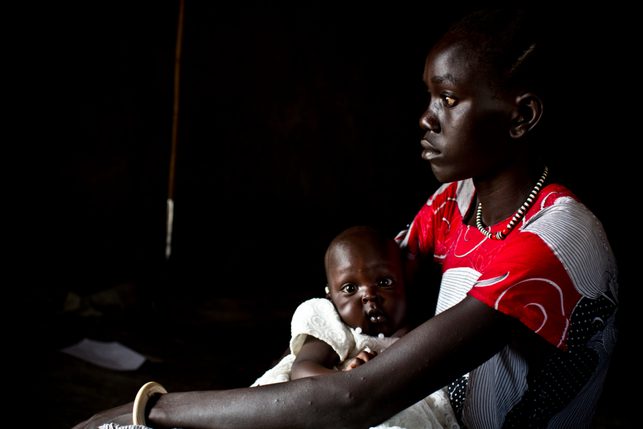A young girl in South Sudan sits with a baby. The girl looks off to the side of the camera, with a sad look on her face. The baby stares into the camera, wearing a white dress.