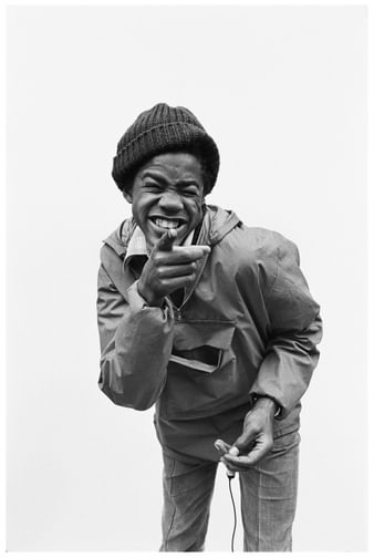 Young Black boy wearing a hat smiles and points at the camera in a black and white photograph