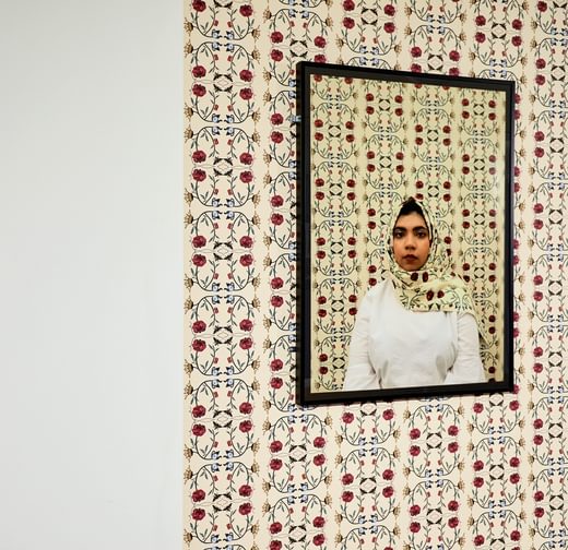 A multi-cultural British person with South Asian heritage is photographed looking into the camera with a patterned headscarf. The same pattern is used as wallpaper in the background. The picture is framed and the wallpaper extends beyond the frame.