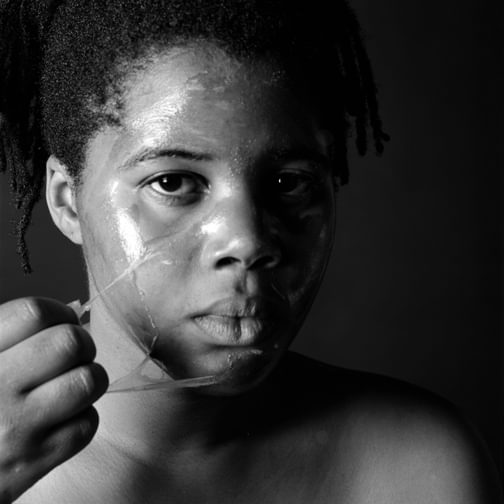Black and white photograph of artist Maxine Walker, looking into camera, peeling something from her facial skin.