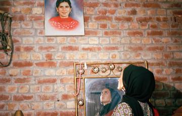Young woman in black hijab looks at a colour painting of her grandmother on a brick wall, the young woman's reflection and face is visible in a mirror