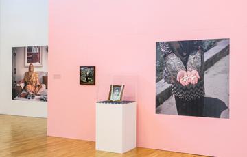 Pink wall and white wall in exhibition with large photographs. Woman with her hands outstretched, woman in saree and hijab. Also a framed photo on the wall and framed photo with flower garland on a plinth.