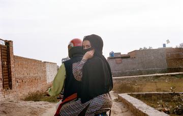 Woman in hijab and saree looks at the camera, face covered, sat on a moped riding along a dusty track in Pakistan