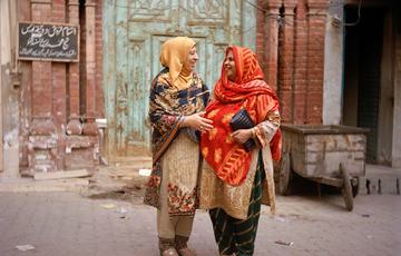 Two women in saree and hijab linking arms, stood in front of a Pakistani building