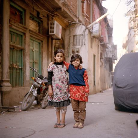 Two children stand on a street in Pakistan in colourful clothes, smiling at the camera
