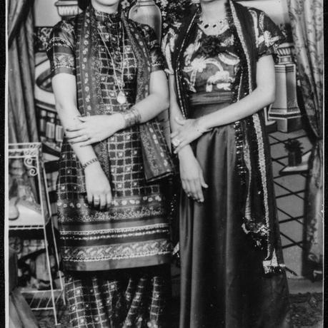 Two women in a black and white archive photo stand in saree smiling at the camera