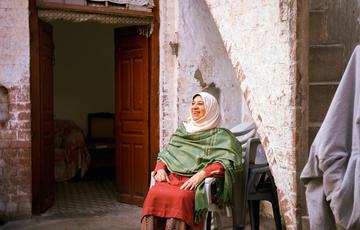 Older woman in saree and hijab (red, green and white) sits on a chair outside of a Pakistani building