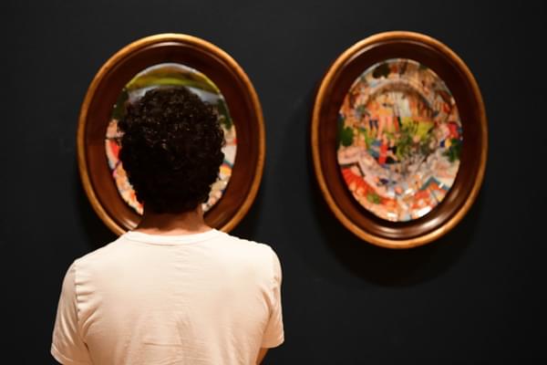 Person faces away from the camera with short black curly hair, looking at artworks. Artworks are painted plates on wooden frame on a dark blue wall