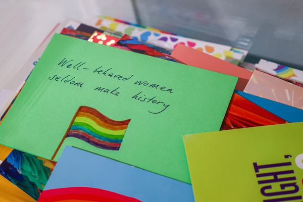 Photograph of a pile of cards. The top card reads 'Well behaved women seldom make history' with a drawing of a rainbow flag.
