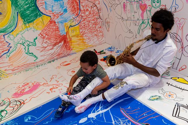 Man playing saxophone with small child drawing on his leg, both smiling. Sat in a gallery with walls covered in colour and drawings.