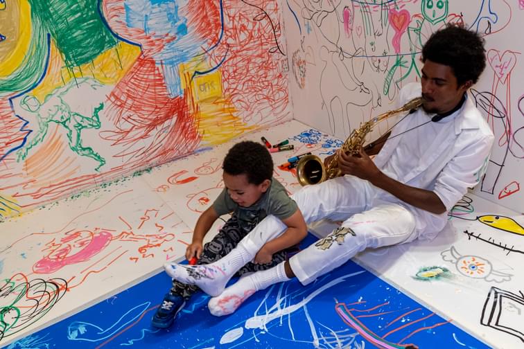 Man playing saxophone with small child drawing on his leg, both smiling. Sat in a gallery with walls covered in colour and drawings.