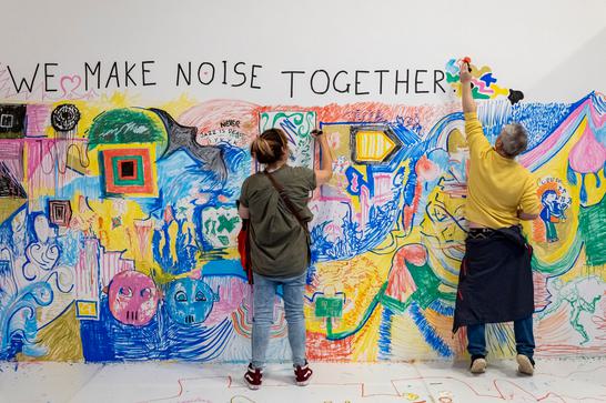 A man and a woman draw on a gallery wall covered in colourful drawings and text that reads 'WE MAKE NOISE TOGETHER'.
