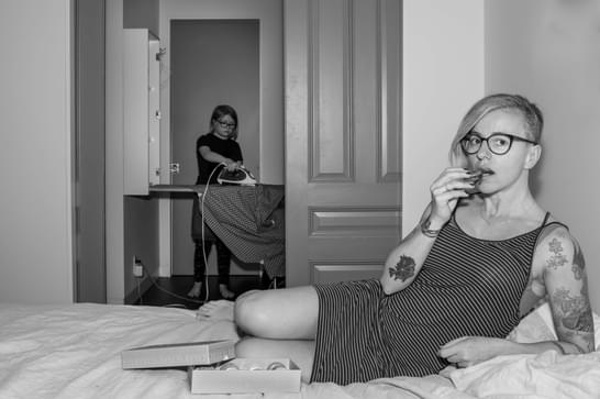 A person sits on a bed eating a macaroon, while a younger child irons in the background