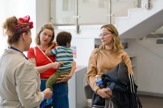 A woman is holding a child in her arms, next to another blonde woman. They are both listening to another woman talking