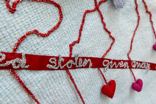 White fabric with red embroidery details, with a red textile strip with the words 'stolen' and 'given away' on top in silver thread