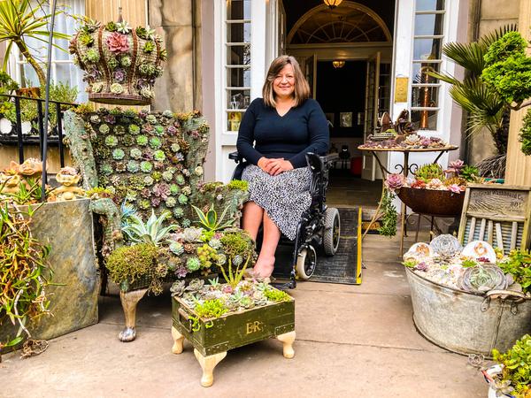 Pauline is to the centre with a front door behind her. To her left there is a throne (chair) that is completely covered in succulents.