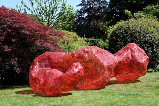 A large red sculpture made from plastic is on grass and surrounded by trees