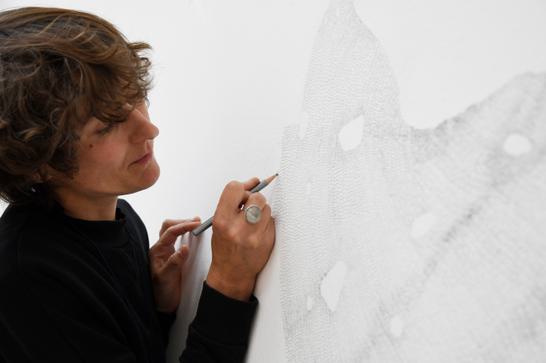 A woman draws with a pencil on a white wall, she has short cropped brown hair and is wearing a black roll-neck jumper. The drawing is an intricate shape with fine lines.