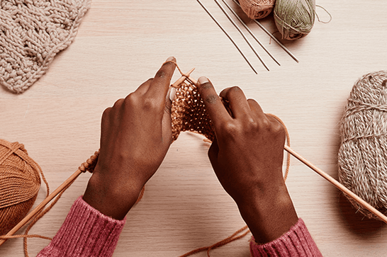 Someone with their hands holding two knitting needles, surrounded by wool
