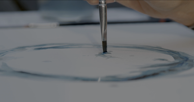 An artist delicately adds darker streaks of watery blue to their circle. At the top its crests like an ocean wave.