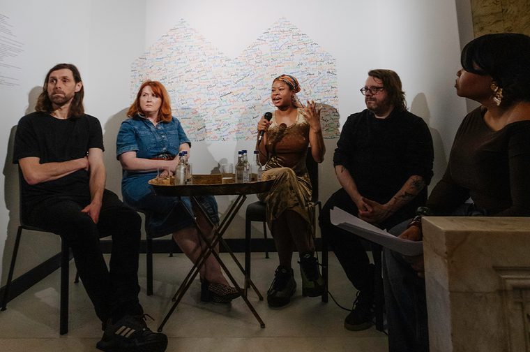 A group of people sat together, one in the centre is holding a microphone, speaking and using expressive hand movements