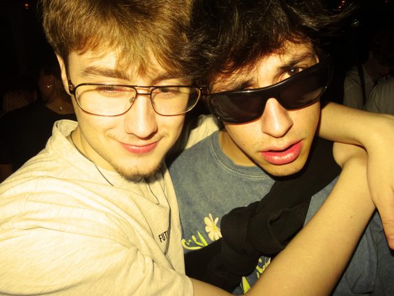 Two people are hugging in a dark room, one is wearing glasses and the other is wearing sunglasses