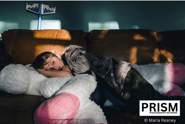 A white boy with dark blond hair (wearing green tracksuit bottoms and a grey long-sleeved T-shirt) snuggles against a large horse or unicorn plushie. He is lying on an tan leather sofa in a dark, shadowy living room. The living room wall behind him is an aquamarine blue.