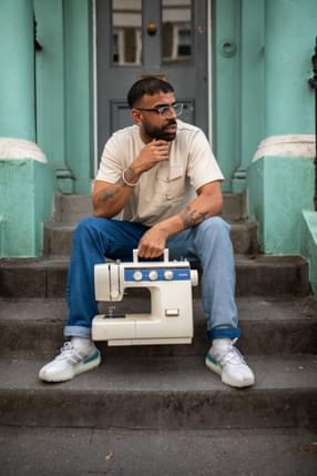 A person is sitting on concrete steps, holding a sewing machine between their legs with one hand. The walls behind them are a light blue.