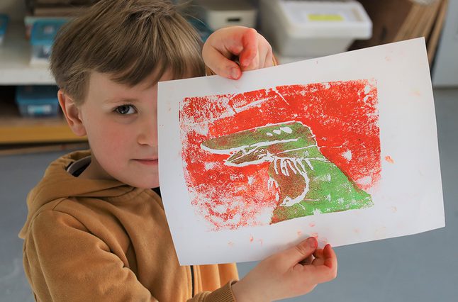 A small child is holding up a piece of paper, with a green dinosaur on and orange background using printmaking techniques