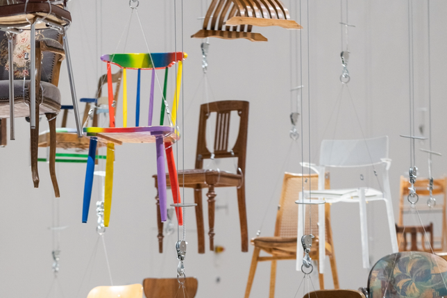 A variety of chairs of different designs and colours are hanging from a ceiling, suspended by silver hoists