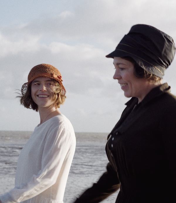 Two smiling women (Jessie Buckley and Olivia Colman) walk along a windswept beach.