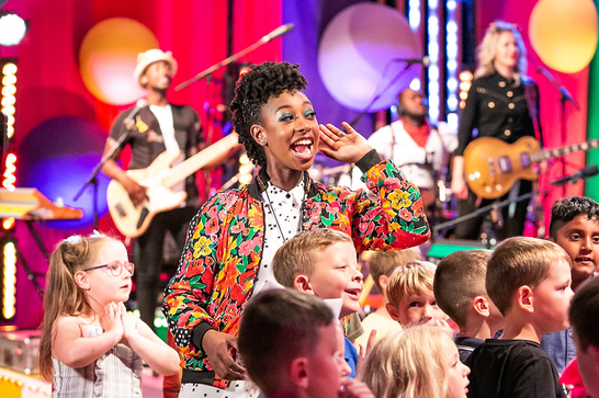 YolanDa Brown holds up a hand to their ear and wears a brightly coloured jacket - there are lots of children in front of them and a band in the background.